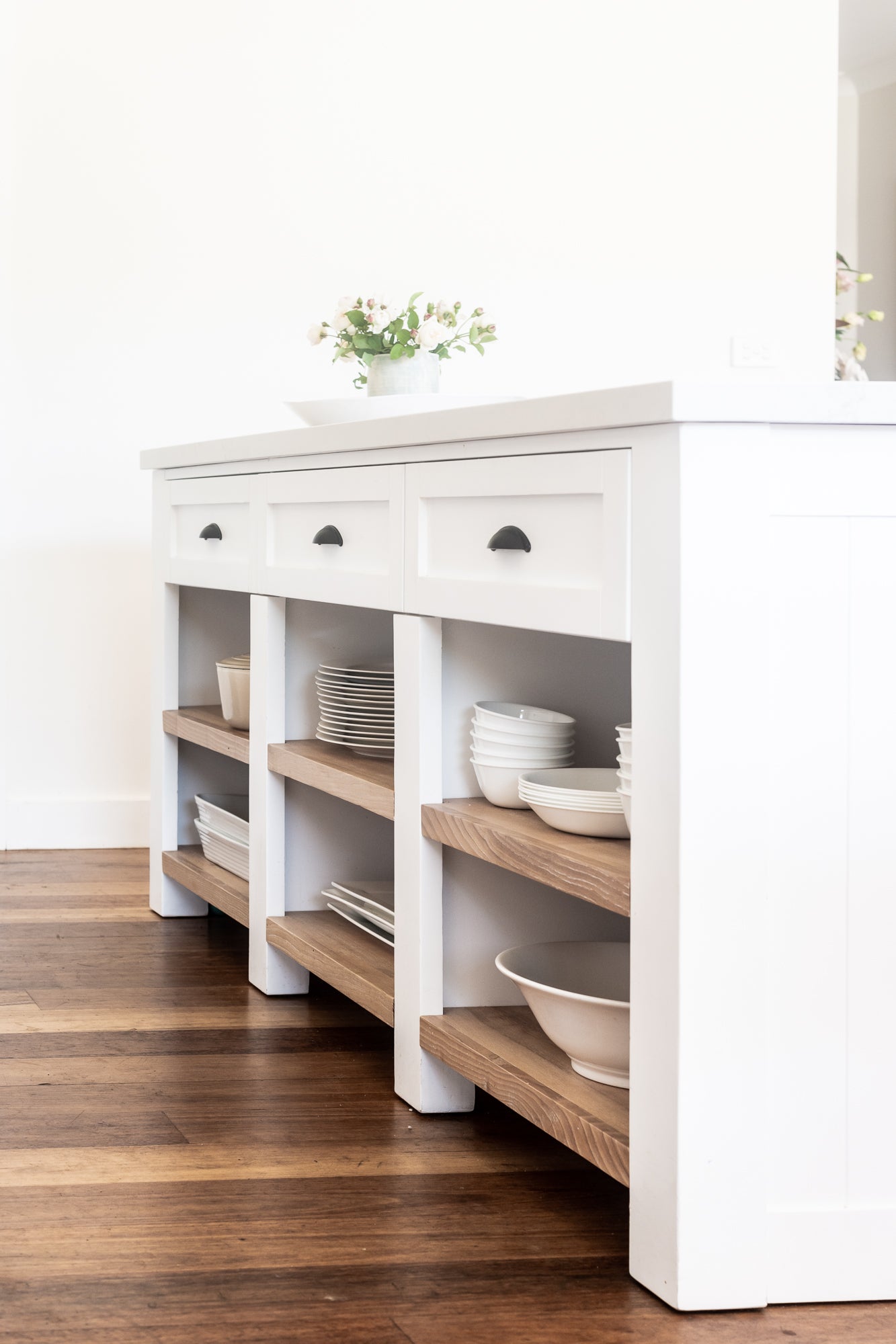 White kitchen island with wooden shelves and various dishes on a wooden floor.