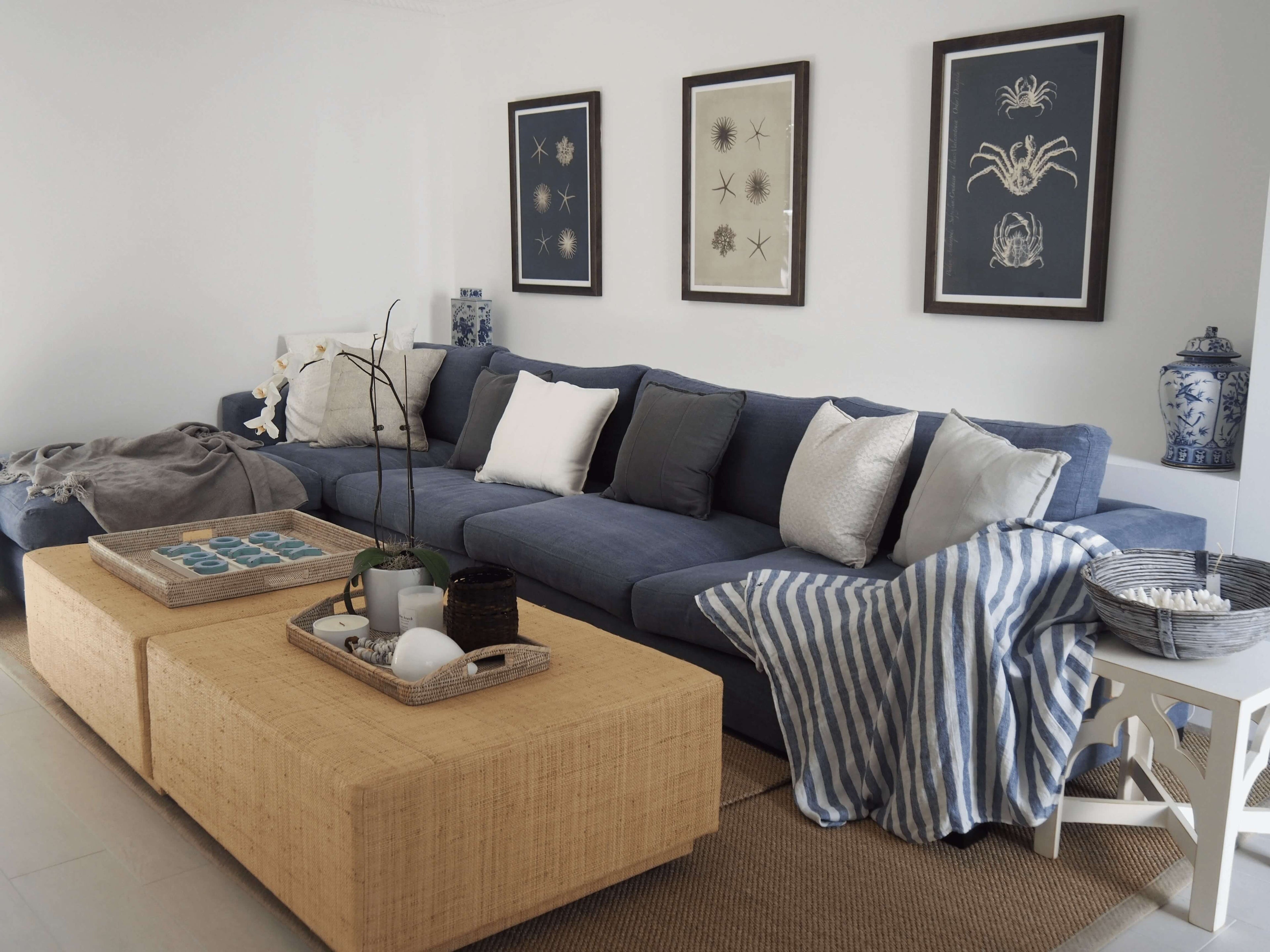 Cozy coastal living room with a custom long blue sofa adorned with white and grey pillows, a natural textured ottoman coffee table, nautical-inspired decor, and botanical framed art on the wall