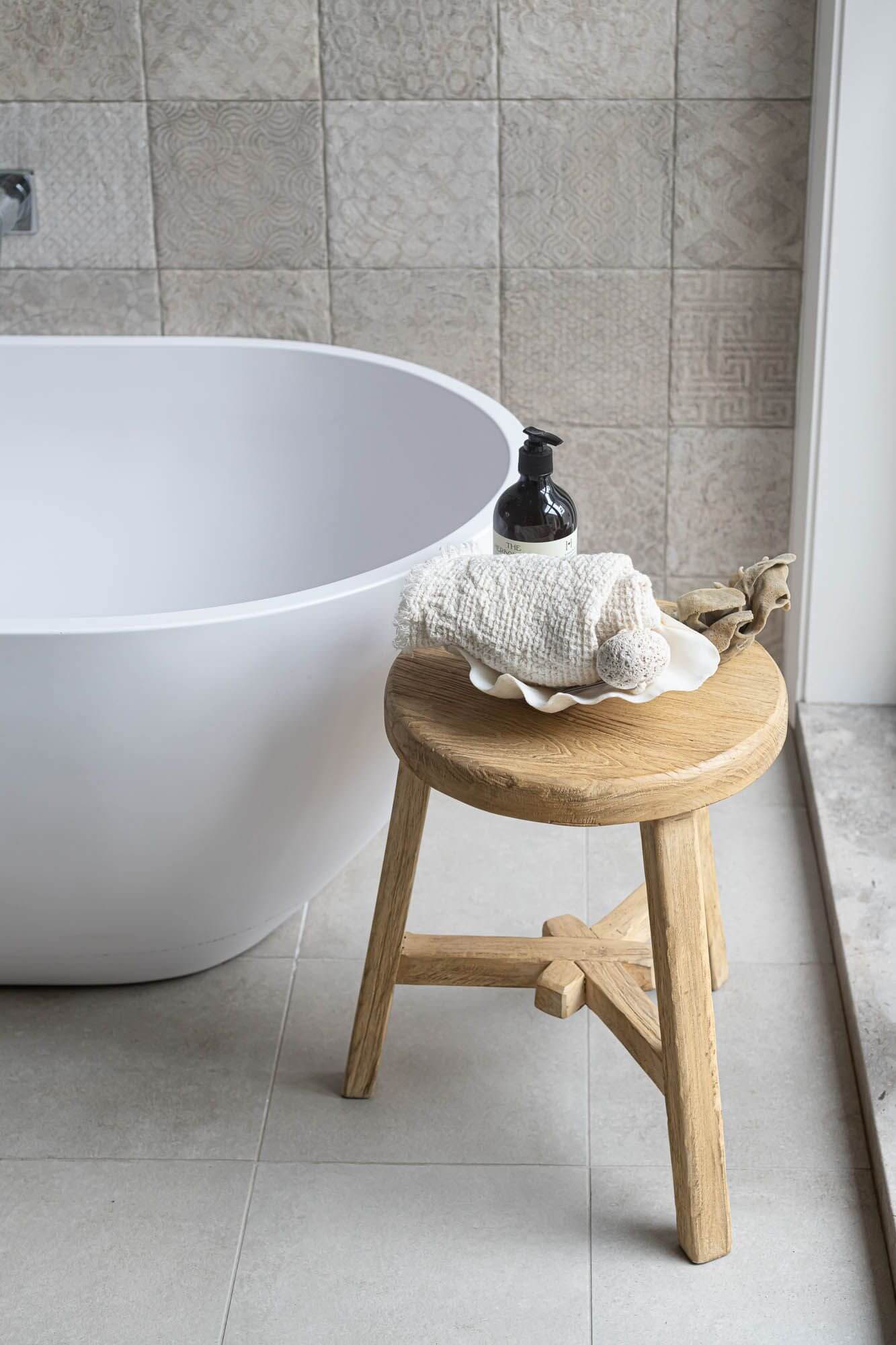 White freestanding bathtub with a wooden elm stool and shell decoration in a tiled bathroom.