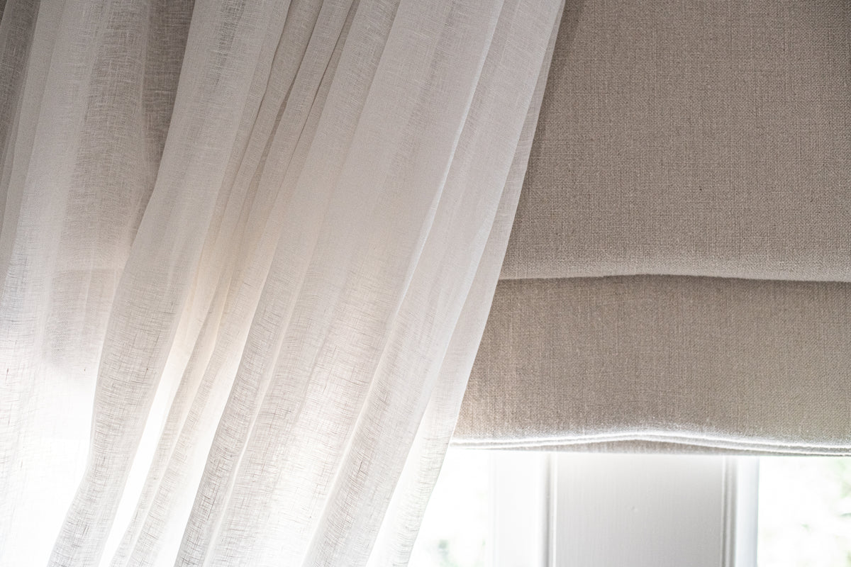 Sheer white curtain with a beige roman blind in front of a window.