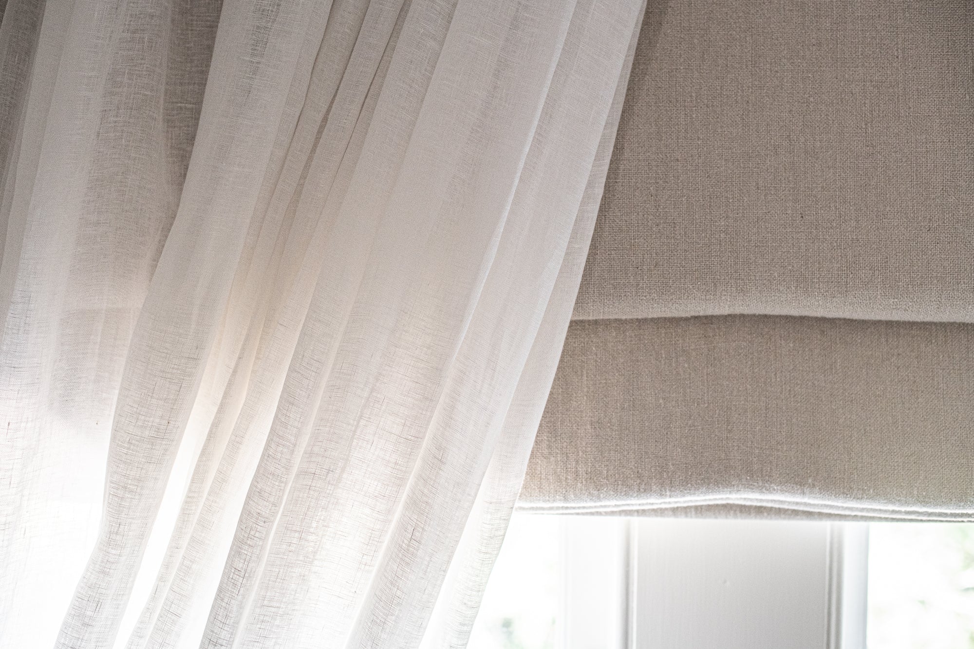 Sheer white curtain with a beige roman blind in front of a window.