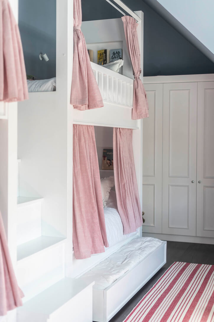 Custom bunk bed with pink curtains in a room with white cabinets and a striped rug.