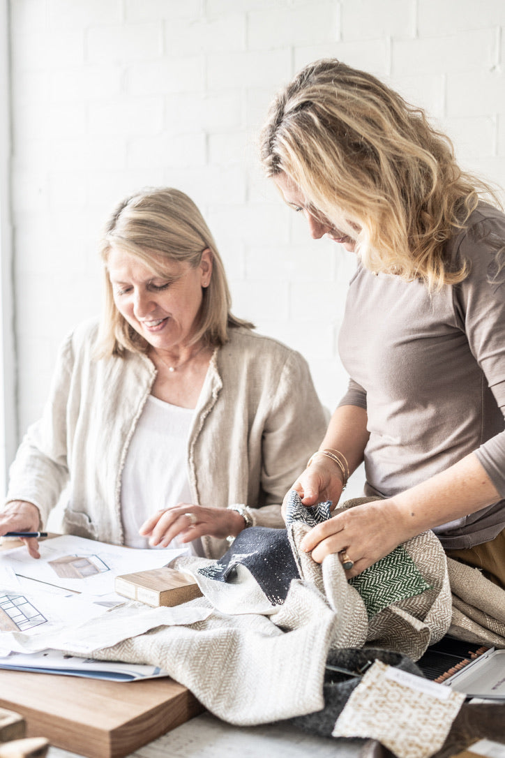 Two women collaborating in a bright, modern room, examining fabric swatches and papers on a wooden table. One woman holds a textured cream fabric while the other looks on.