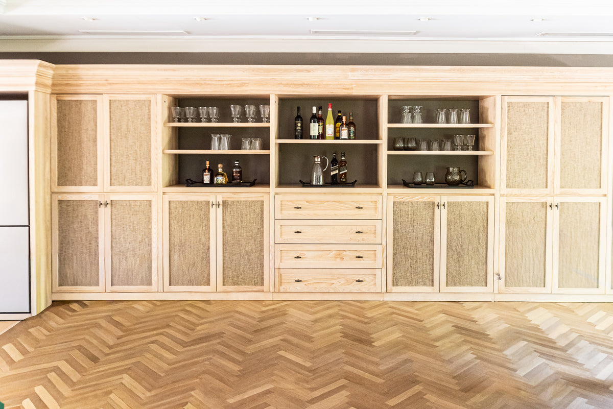 A large built-in wooden bar cabinet with woven-style doors, open shelves holding glassware and bottles, and a central set of drawers, shown in a bright interior with wood herringbone flooring.