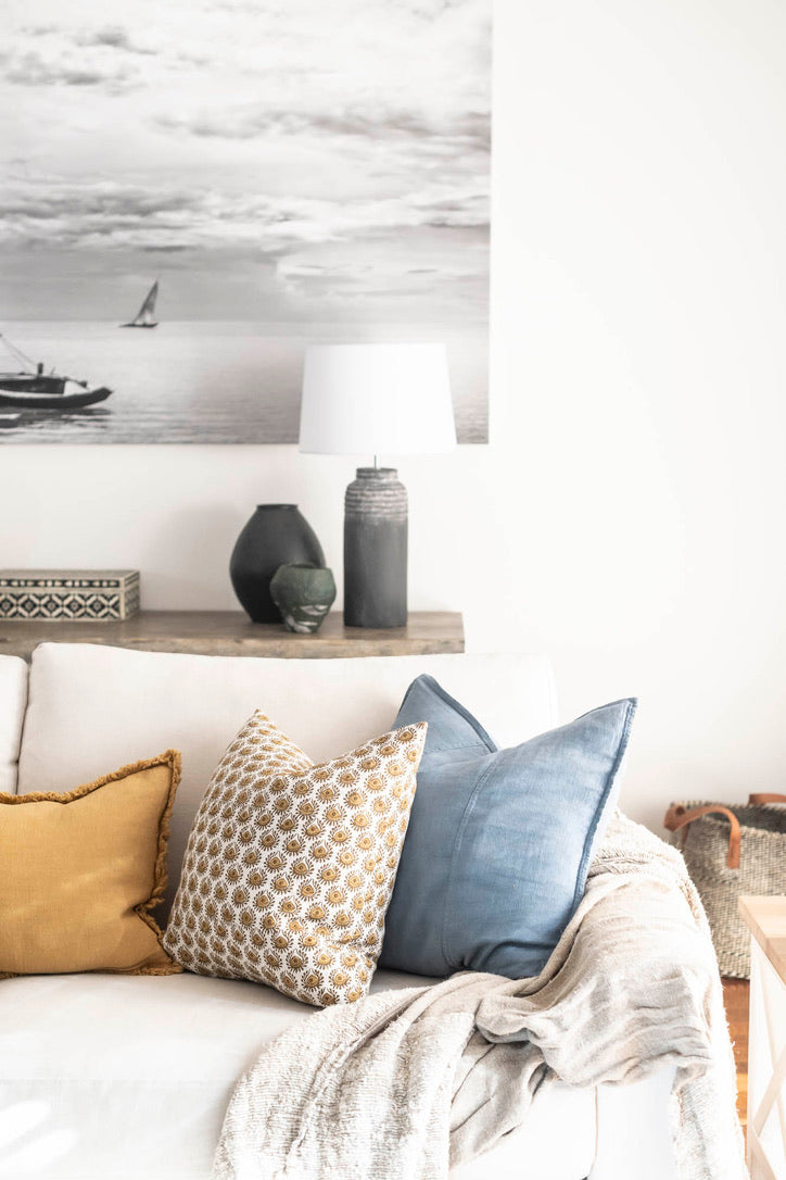 A styled living room corner featuring a white sofa with decorative throw pillows in mustard yellow, beige-and-gold patterned, and dusty blue, with a cream throw blanket. A floating shelf holds a lamp and decorative vases, and a large black-and-white nautical art print hangs on the wall above.