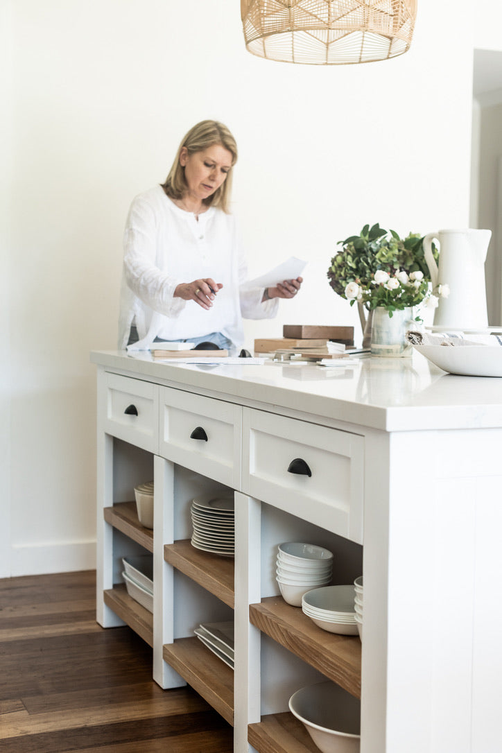 Woman in a kitchen preparing food at a white island with wooden shelves.