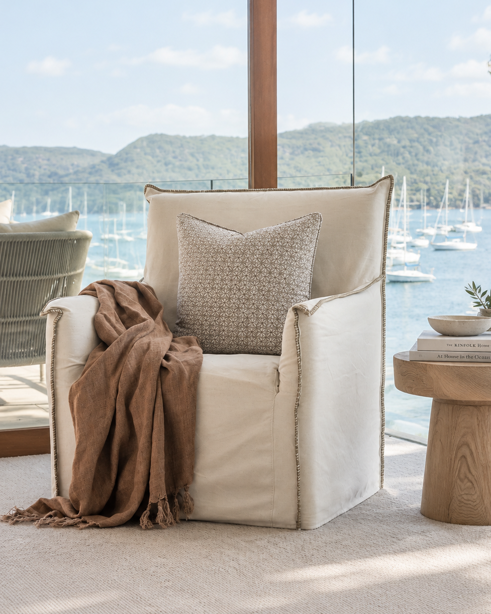 A cream upholstered armchair with a textured beige throw pillow and a draped brown throw blanket, styled next to a round wooden side table in a bright room overlooking a scenic lake and marina view.