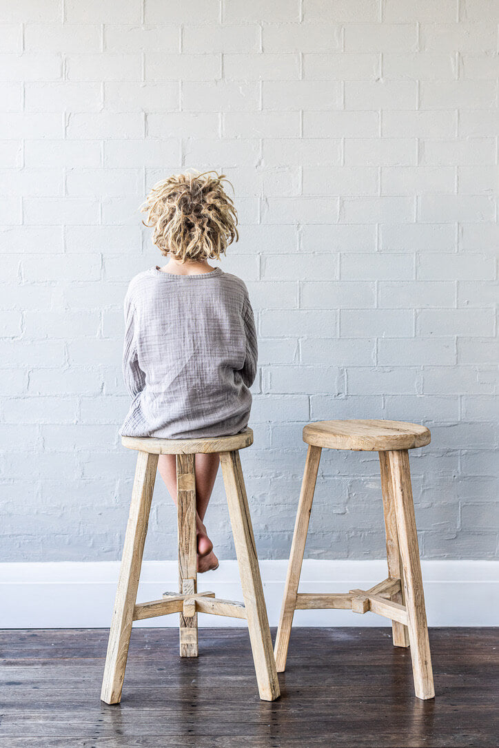 Person sitting on a wooden elm stool in front of a grey brick wall