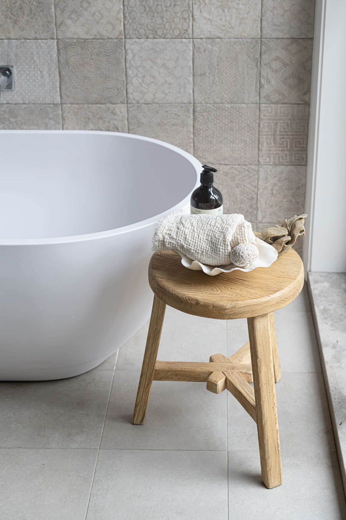 White freestanding bathtub with a wooden elm stool and shell decoration in a tiled bathroom.