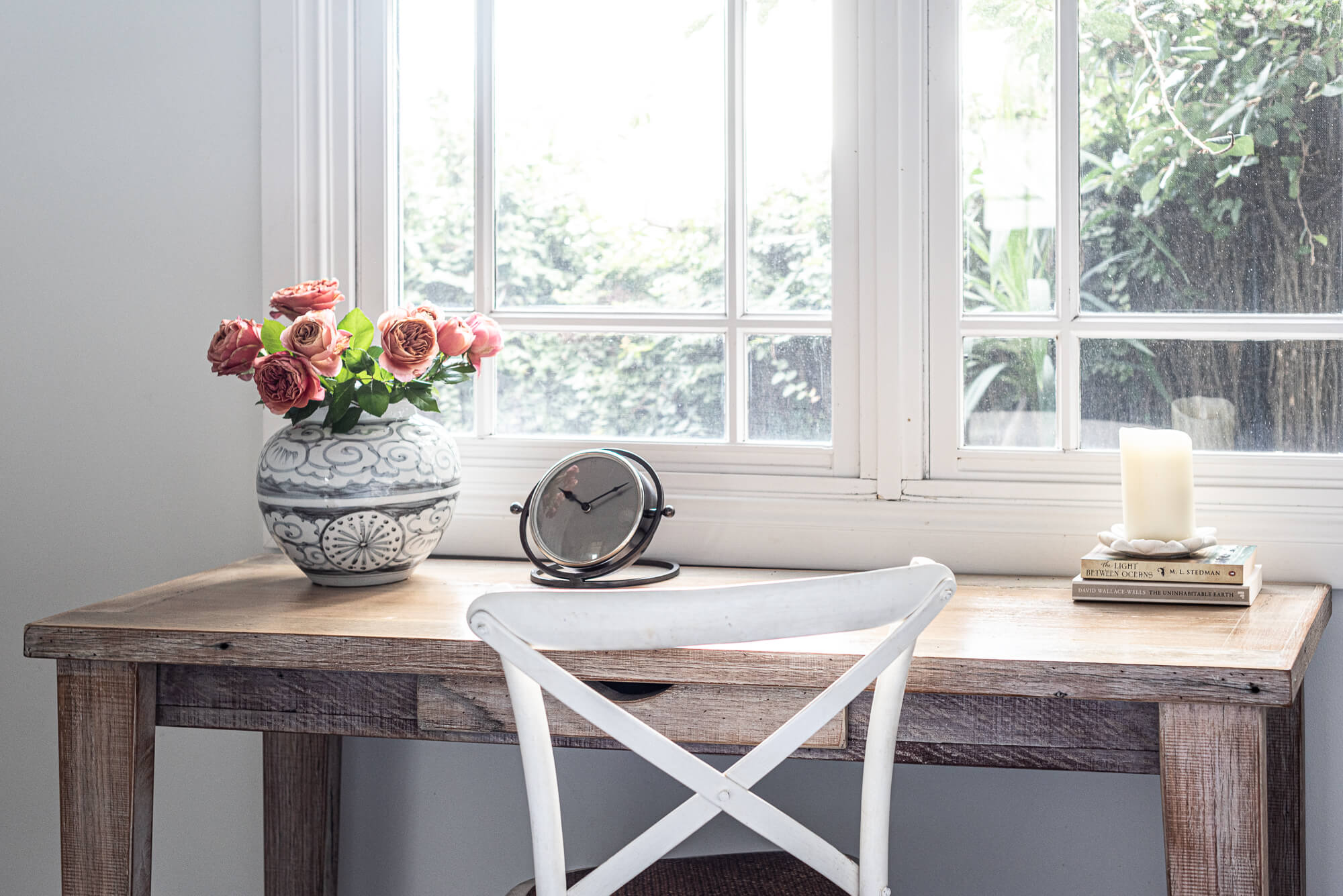 Wooden desk with a vase of flowers, clock, and candle in front of a window.