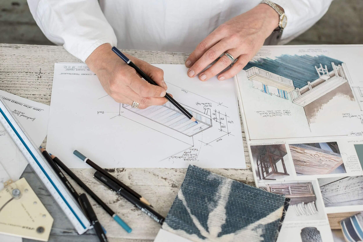Interior designer drafting design sketches with pencil and ruler, with fabric samples and inspiration images spread out on a worktable.
