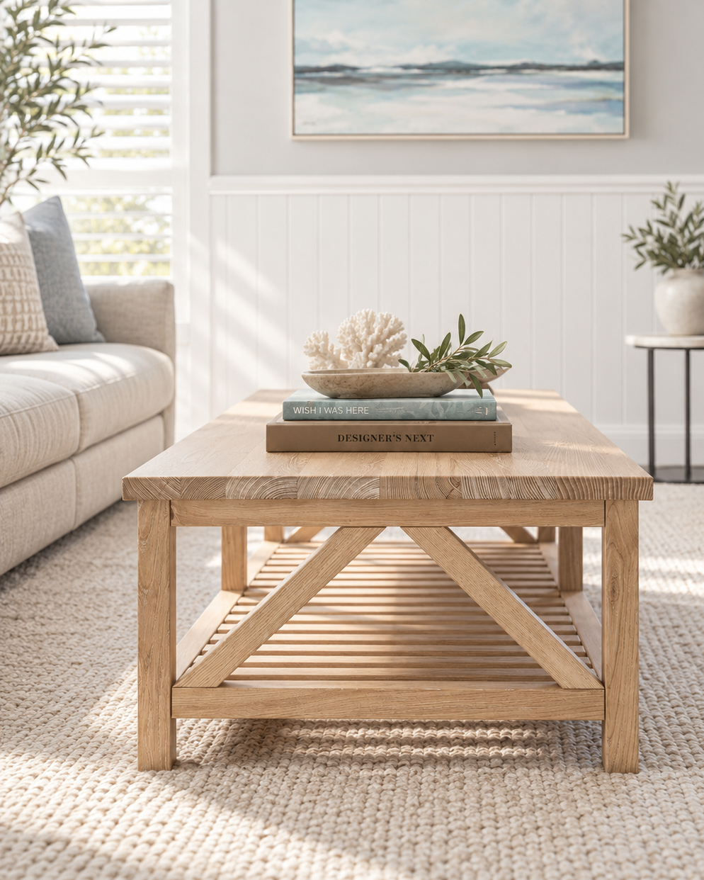 A light natural wood coffee table with a slatted lower shelf, styled in a bright, modern living room with a beige sofa, a striped armchair, and a framed ocean painting on the wall.